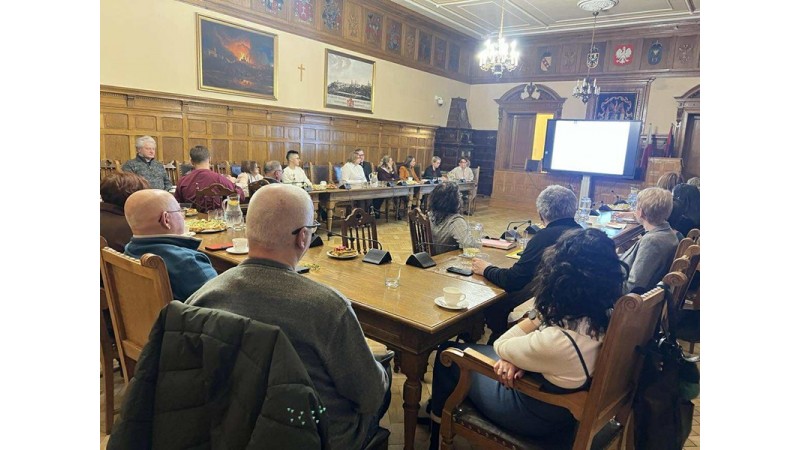 Spotkanie Noworoczne z udziałem przedstawicieli organizacji pozarządowych w Sali Sesyjnej cieszyńskiego Ratusza. Duża, zabytkowa sala. Przy długim stole siedzi kilkanaście osób i patrzy na ekran z prezentacją. Na stole są mikrofony, woda i filiżanki. 